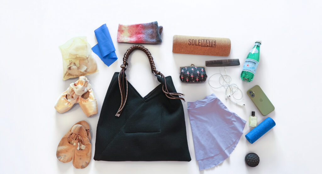 On a white background are a black bag and several items including a purple skirt, pointe shoes and ballet shoes, a band and half foam roller, and a phone with corded headphones