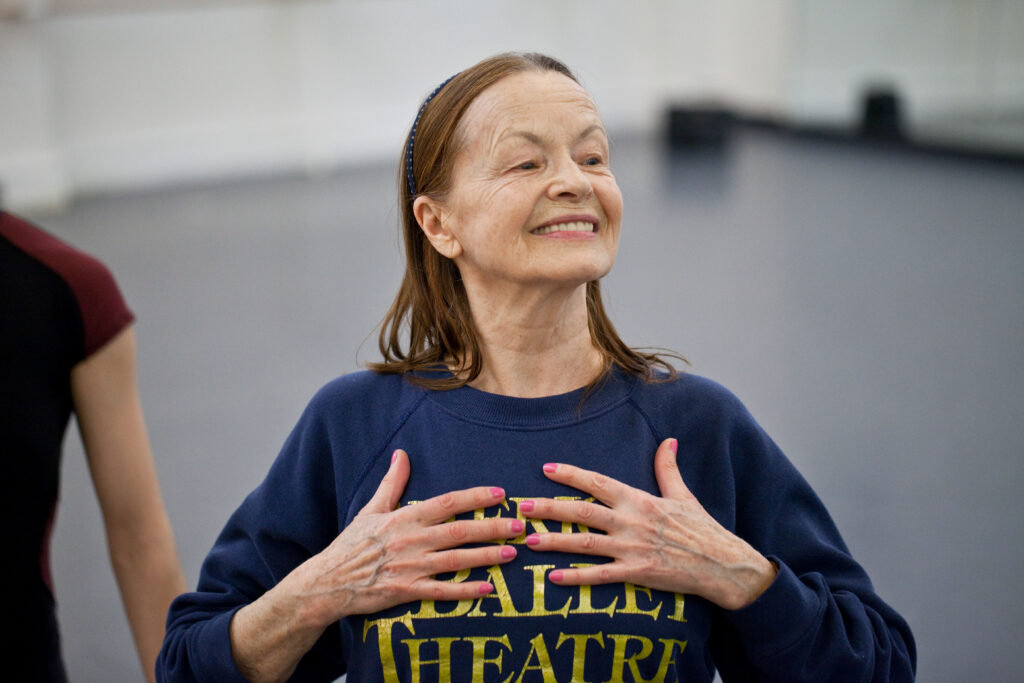 Irina Kolpakova stands wearing a navy ABT sweatshirt. She smiles and gestures toward herself with her hands.