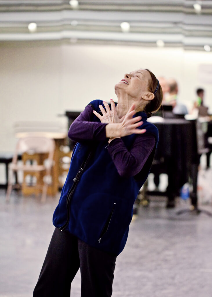 Irina Kolpakova dramatically leans back with her arms folded across her chest in a brightly lit studio. She is wearing a navy vest, purple shirt, and black pants.