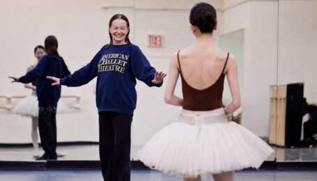 In a rehearsal, Irina Kolpakova smiles at a dancer and stands with her arms placed out to the side. The dancer wears a maroon leotard and a white tutu and looks toward her.