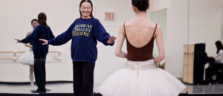 In a rehearsal, Irina Kolpakova smiles at a dancer and stands with her arms placed out to the side. The dancer wears a maroon leotard and a white tutu and looks toward her.