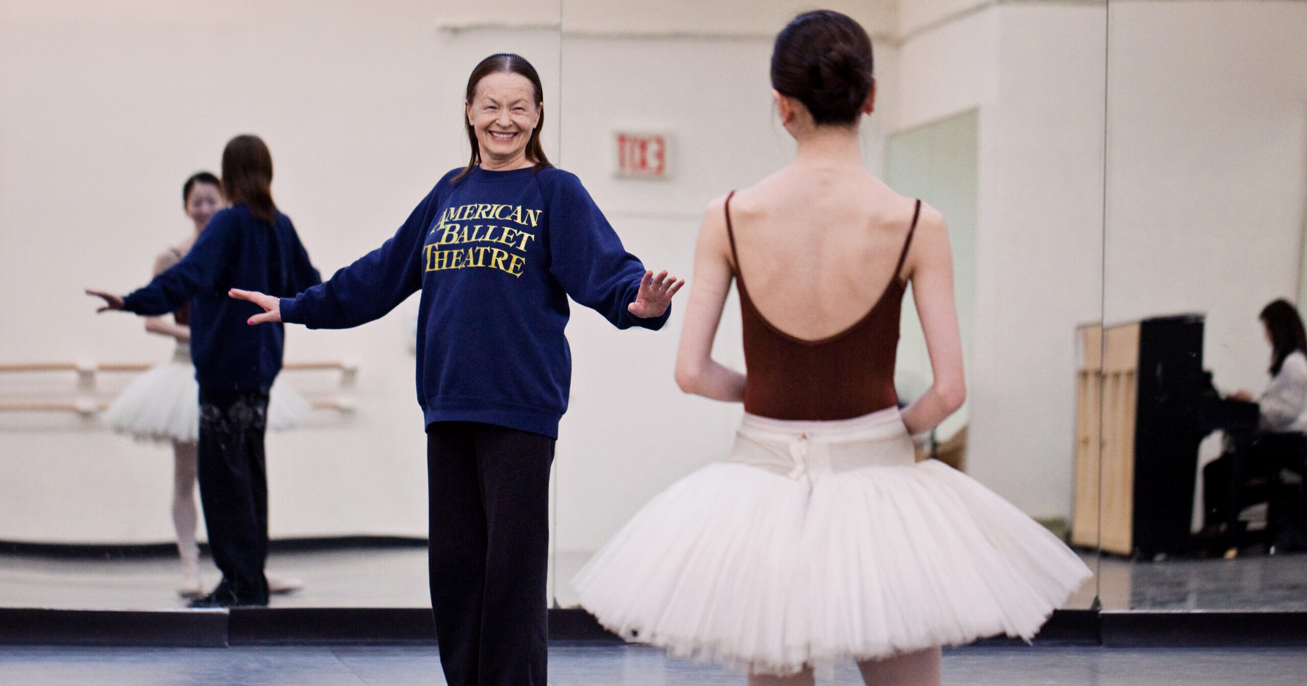In a rehearsal, Irina Kolpakova smiles at a dancer and stands with her arms placed out to the side. The dancer wears a maroon leotard and a white tutu and looks toward her.