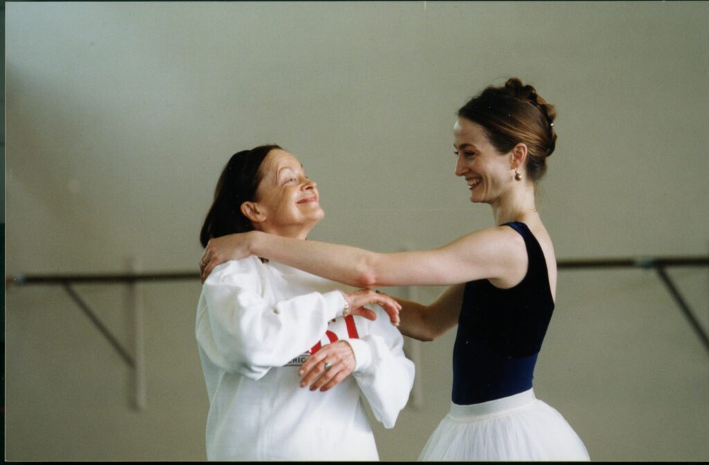 In a studio, Irina Kolpakova faces Julie Kent, smiling. Kent has her left arm outstretched toward Kolpakova. Kent is wearing a navy leotard and a white tutu, and Kolpakova is wearing a white sweatshirt.