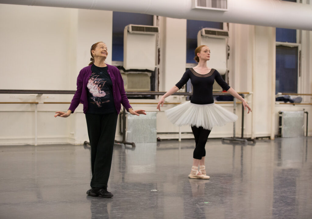 In a studio, Irina Kolpakova stands smiling in fifth position with her arms at her sides, wearing black pants, a purple cardigan, and a black t-shirt featuring dancers. Next to her, Gillan Murphy stands in fifth position wearing black tights, a black leotard, and a white tutu.