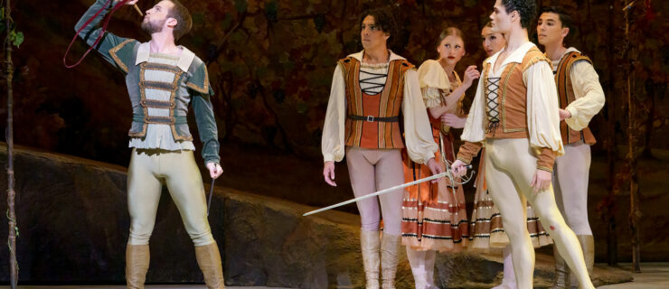 Carraig New blows a horn while Ángel Ramírez stands next to him with a sword. Corps dancers look on behind them. They wear medieval style costumes.
