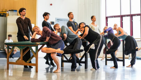 In a brightly lit studio, nine dancers rehearse surrounding a long table. On one side, five of them lean one elbow on the table and cross one leg over the other. On the other side, four dancers stand stiffly with both arms reaching forward toward the table.
