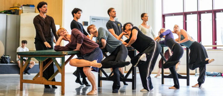 In a brightly lit studio, nine dancers rehearse surrounding a long table. On one side, five of them lean one elbow on the table and cross one leg over the other. On the other side, four dancers stand stiffly with both arms reaching forward toward the table.
