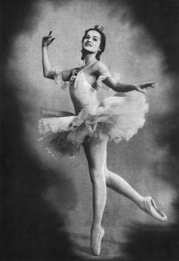 Irina Kolpakova poses on pointe with her right leg slightly elevated to the back. Her left arm is stretched to the side, and her right is to the side with the forearm bent so her palm faces toward her. She looks up and back, wearing a tutu, and smiling. The image appears in black and white.