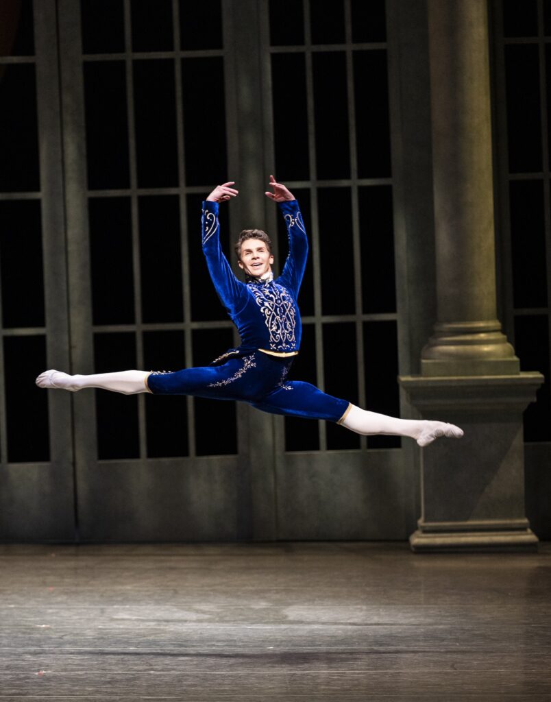 On stage, a male dancer in a blue-and-gold jacket, shorts, white tights, and ballet slippers, does a saut de chat with his arms up.