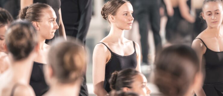 Dancers wearing black leotards and black skirts stand attentively in a studio.