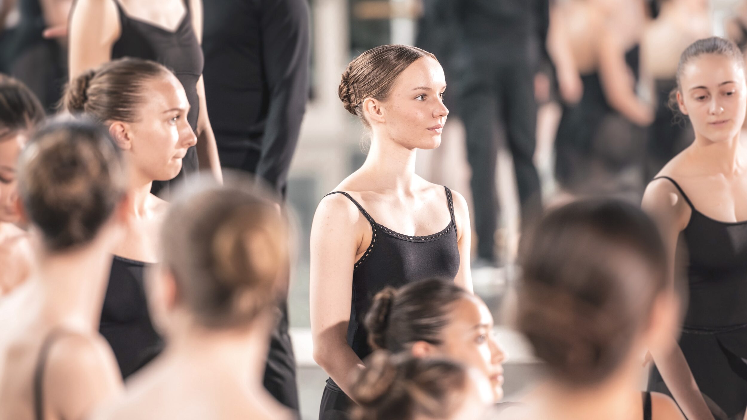 Dancers wearing black leotards and black skirts stand attentively in a studio.