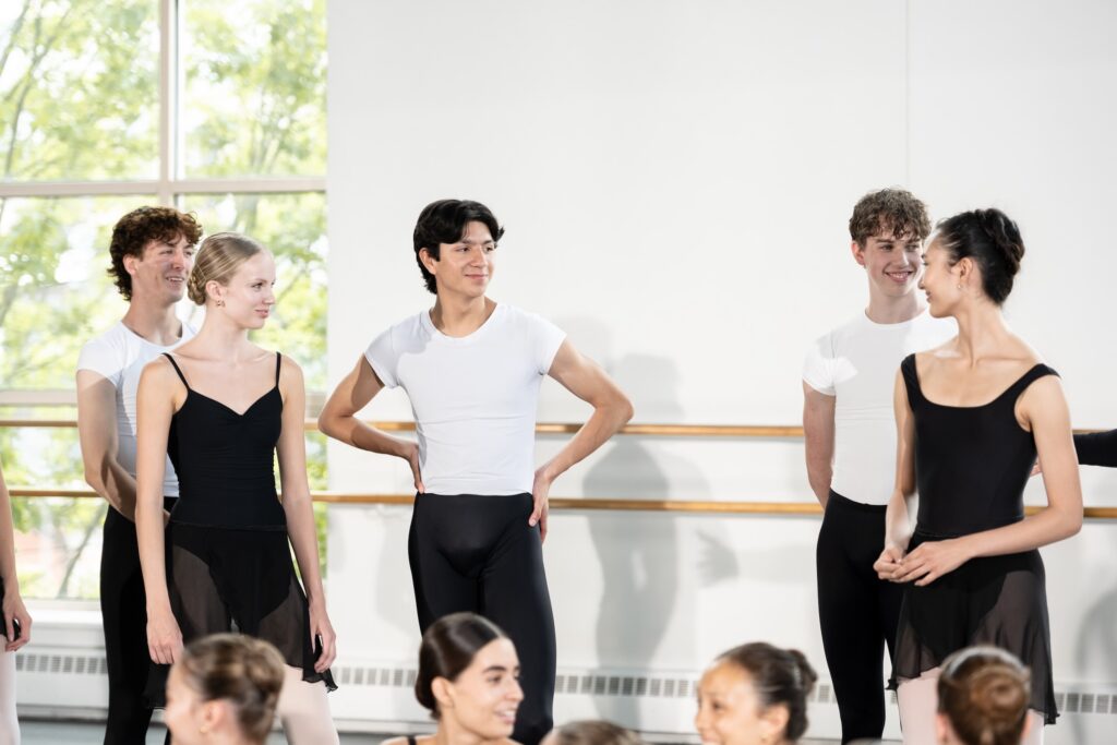 In a dance studio, two dancers wearing black leotards and black skirts stand smiling at each other. Behind them, three dancers wearing white shirts and black tights smile as they interact with the pair.