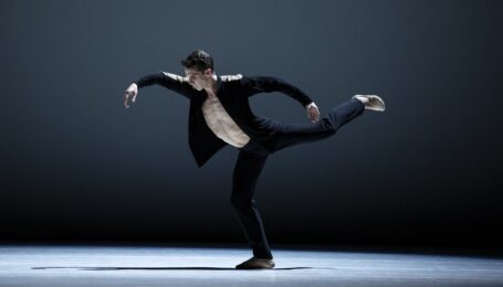 On a stage with a grey background, one male dancer appears in a black jacket, pants, and beige ballet slippers. He has his left leg elevated in an attitude with the foot flexed. His arms are elevated and curved.