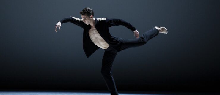 On a stage with a grey background, one male dancer appears in a black jacket, pants, and beige ballet slippers. He has his left leg elevated in an attitude with the foot flexed. His arms are elevated and curved.