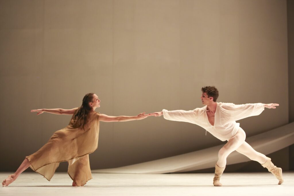 Two dancers appear on stage with a beige backdrop. The female dancer wears a dark beige tunic and poses in a lunge with her right leg tendu back. Both dancers' right arms reach toward the other, just enough to allow their fingertips to touch. The male dancer wears a white tunic, tights, and boots while posing in a lunge with his right leg tendu back.