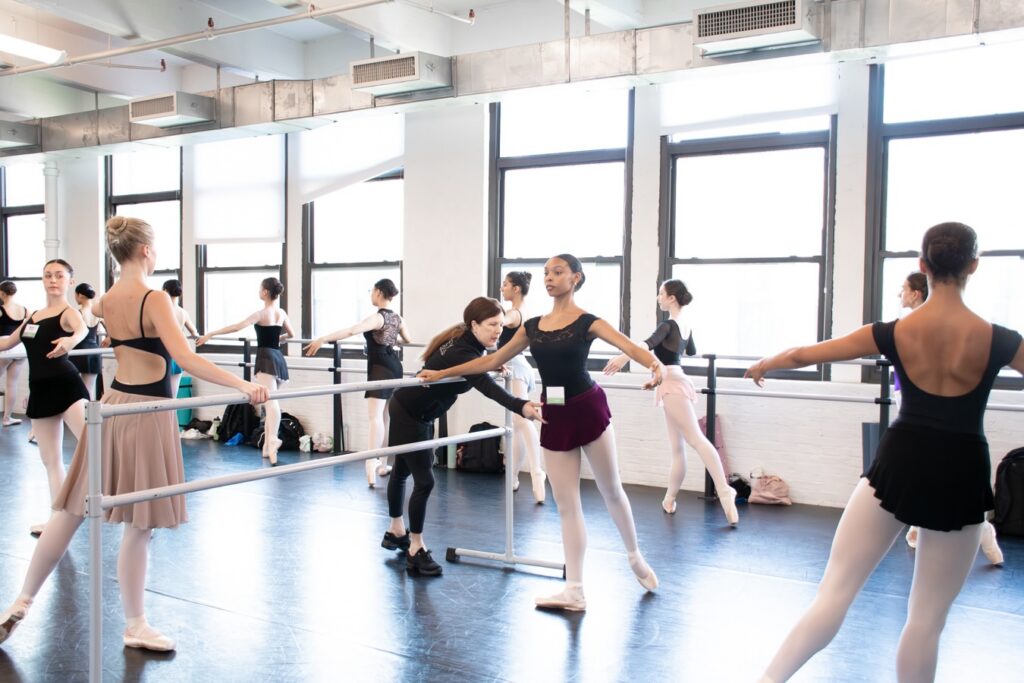 Many dancers wearing black leotards and skirts stand at the barre. In the center, a dancer with her leg extended behind her in tendu receives a correction from her instructor.