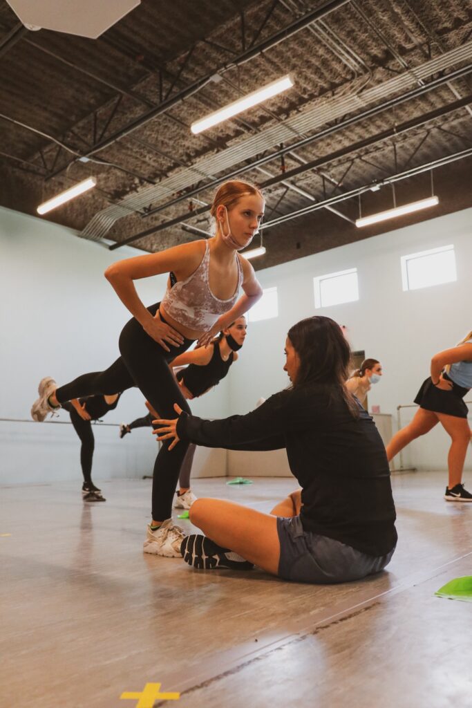 Danielle Farzanegan sits on the floor and helps stabilize a young dancer's knee as she tries to balance in arabesque plié with her hands on her hip. The dancer wears black leggings, leotard, and sneakers. Other dancers in the studio are practicing the same exercise.