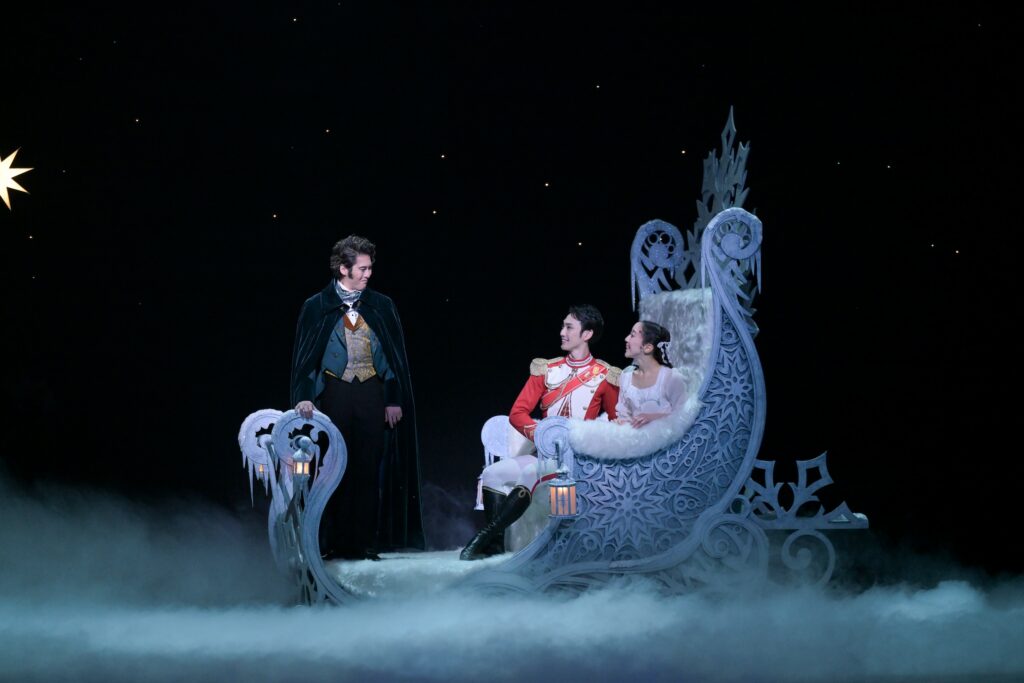 Onstage, two dancers sit in an ice-covered sleigh. The female is wearing a white dress, and the male is wearing a red, white, and gold-adorned jacket. A third dancer wears a green cape and smiles down at them.