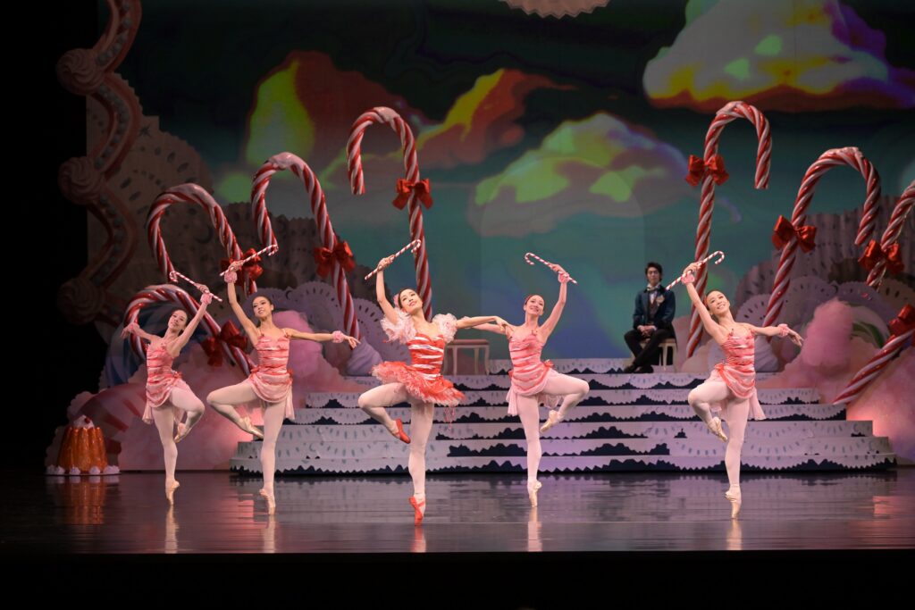 Onstage, four dancers wear red-and-white striped short dresses. In the center, a dancer is wearing a red-and-white striped tutu and red pointe shoes. Each dancer stands on pointe in passé with an arm raised holding a candy cane, while the other arm is held in second.