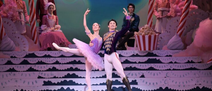 Two dancers pose together on stage. The female dancer wears a lilac tutu and stands on pointe in a back attitude with her right leg elevated and right arm up. The male dancer supports her waist with his right arm, while his other arm is up. The pair smile out toward the audience.
