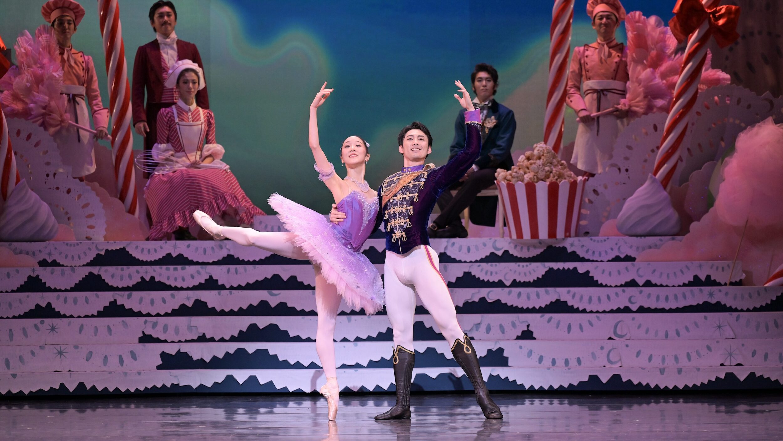Two dancers pose together on stage. The female dancer wears a lilac tutu and stands on pointe in a back attitude with her right leg elevated and right arm up. The male dancer supports her waist with his right arm, while his other arm is up. The pair smile out toward the audience.
