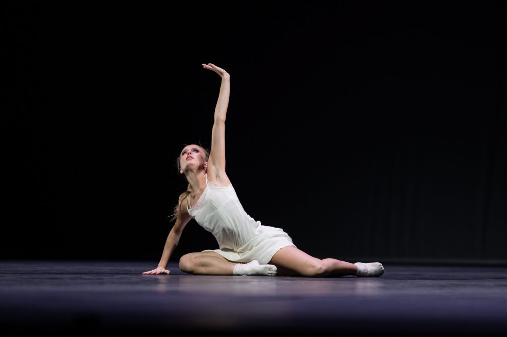 A young female dancers in a white, filmy dress,sits on the floor with her legs slightly tucked and extending to her left. She presses the floor with her right hand and reaches her left arm up, looking skyward.