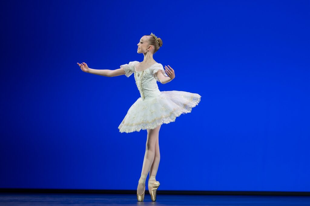 A young teenage ballerina in a white tutu poses in sus-sous on pointe with her feet in croisé. She smiles and looks towards downstage right with her arms opening to the sides, palms up.