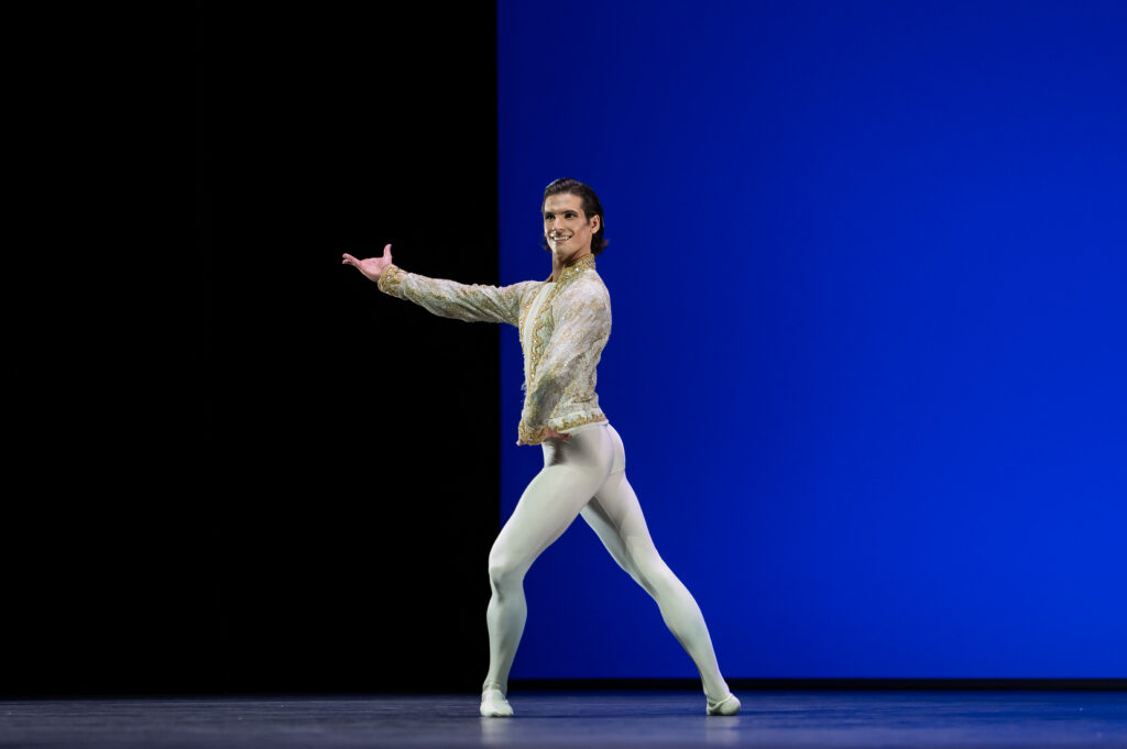 A male ballet dancer poses in a wide fourth position croisé devant with his front leg bent slightly. He places one hand on his hip and extends the other with bravura. He wears white tights and a white tunic with gold details.