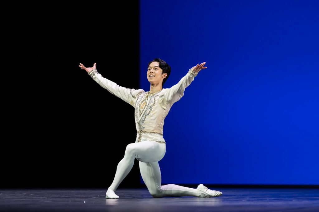 A young male dancer in a white tunic and white tights kneels on his right leg, facing the audience. He lifts his arms up slightly with a triumphant smile.