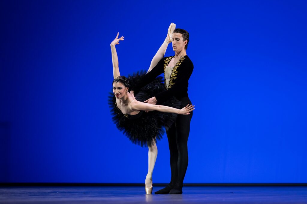 A male and female dancer perform a pas de deux onstage. The woman, wearing a black tutu, does a penché towards the audience, her arms pressed out to the side. Her partner, in a black tunic and lack tights, stands behind her and holds her by the waist.