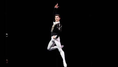 A young male dancer in a black velvet tunic and white tights and ballet slippers jumps up, his right leg in passé. He puts his left hand on his hip and lifts his right arm up, and looks over his left shoulder.