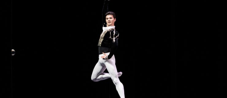 A young male dancer in a black velvet tunic and white tights and ballet slippers jumps up, his right leg in passé. He puts his left hand on his hip and lifts his right arm up, and looks over his left shoulder.