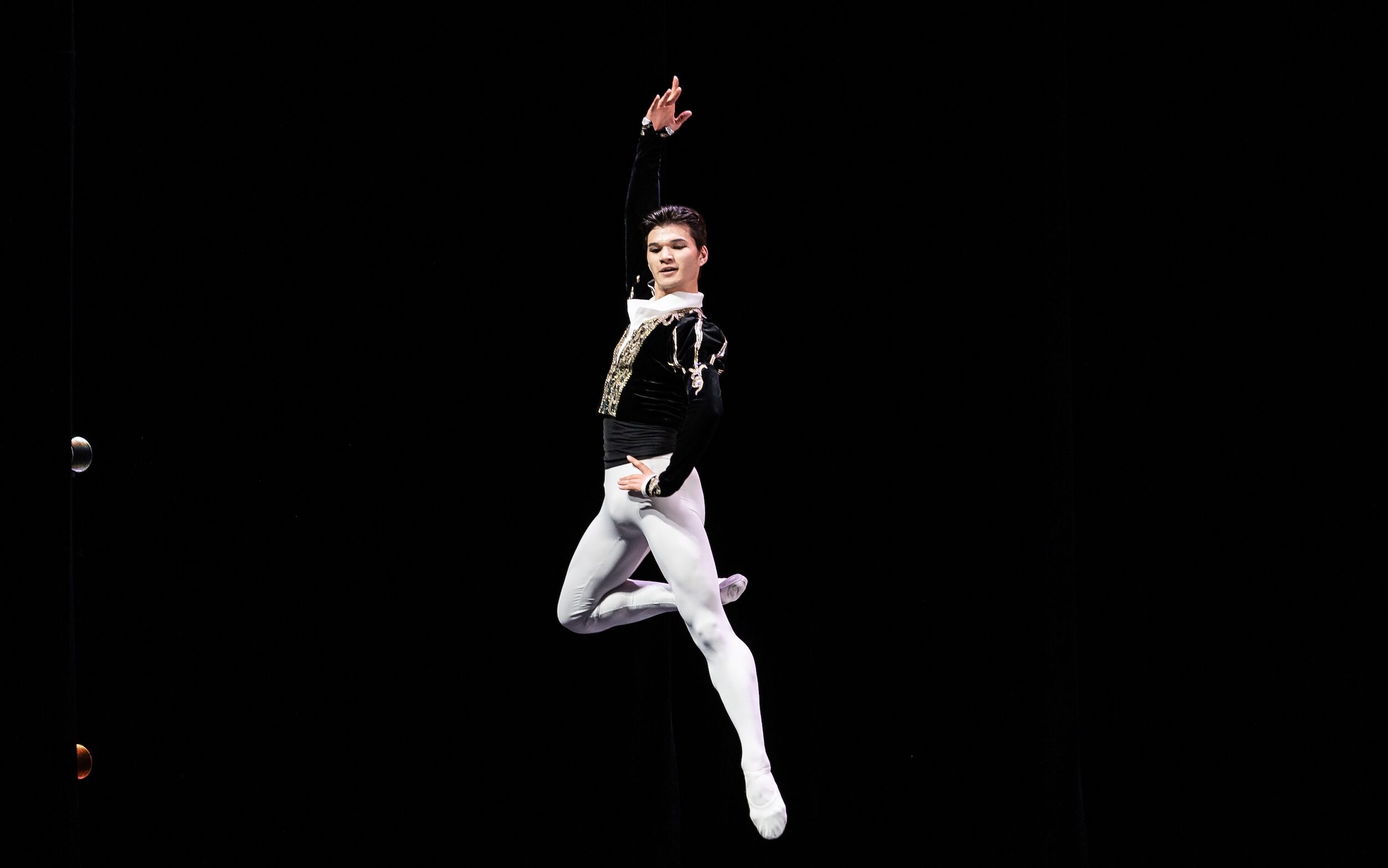 A young male dancer in a black velvet tunic and white tights and ballet slippers jumps up, his right leg in passé. He puts his left hand on his hip and lifts his right arm up, and looks over his left shoulder.