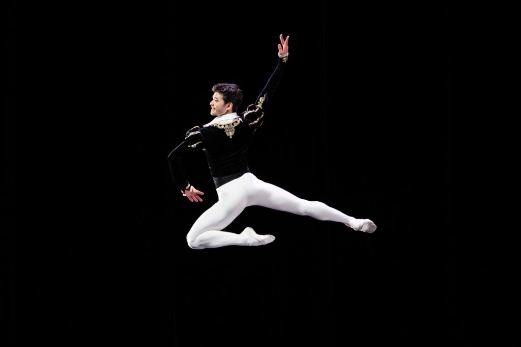 A male dancer jumps up, extending his right leg back and tucking his right leg underneath him. He wears a lack velvet tunic, white tights, and white ballet shoes. He looks over his left shoulder towards the audience.