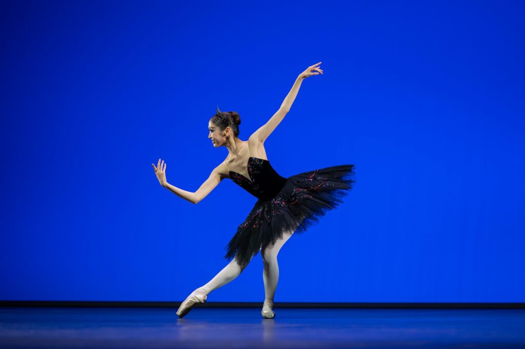 A young ballerina in a black tutu and pointe shoes pliés in tendu with her right leg extended towards stage right. She leans forward slightly as she moves her right arm down and lifts her left arm up behind her.