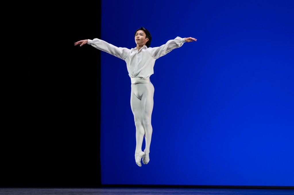 A young male dancer in a white tunic and tights and white ballet slippers jumps up in fifth position with his arms out to the side.