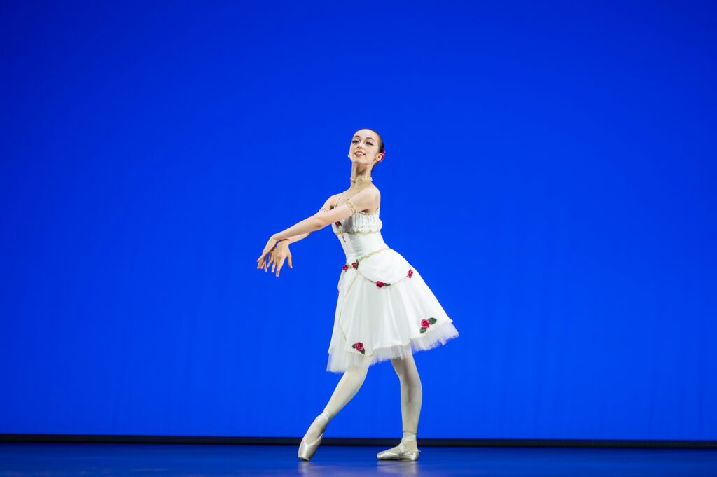 A young ballerina in a knee-length white tutu decorated with rosettes poses in tendu devant croisé. She extends her arms in front of her and crosses her wrists, smiling at the audience.