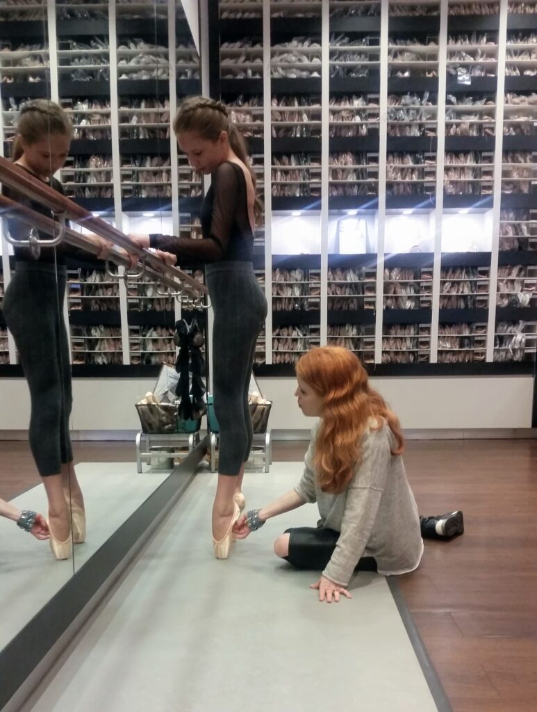 Facing a ballet barre, a dancer wearing a black leotard and dark green leggings stands on pointe in first position. Mary Carpenter sits at her feet and adjusts the dancer's left foot.