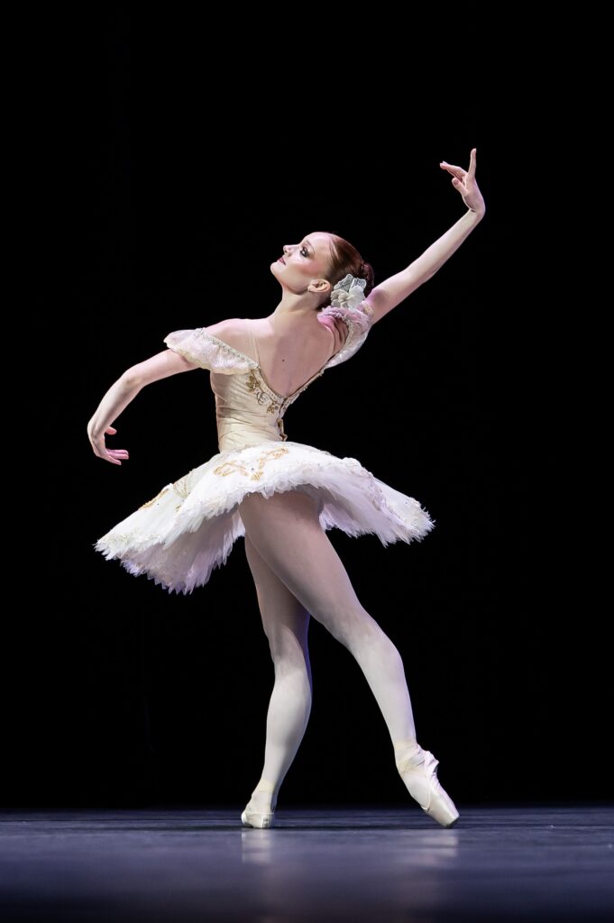 A young ballerina in a white tutu with gold trim poses in tendu derrière. She faces stage right and twists her upper body slightly, with her curved right arm lifted and slightly behind her and her front arm curved low in front.