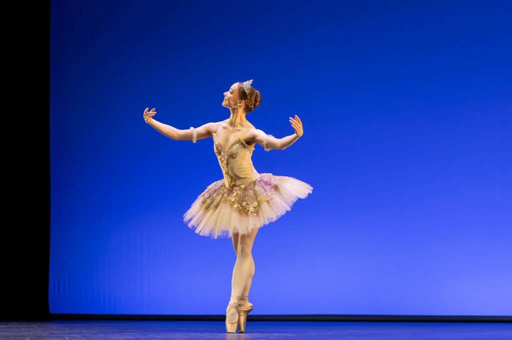 A young teenage ballerina in an offwhite tutu with gold trim and sparkly tiara poses in sus-sous on pointe with her feet in croisé. She smiles and looks towards downstage right with her arms opening to the sides, palms up.