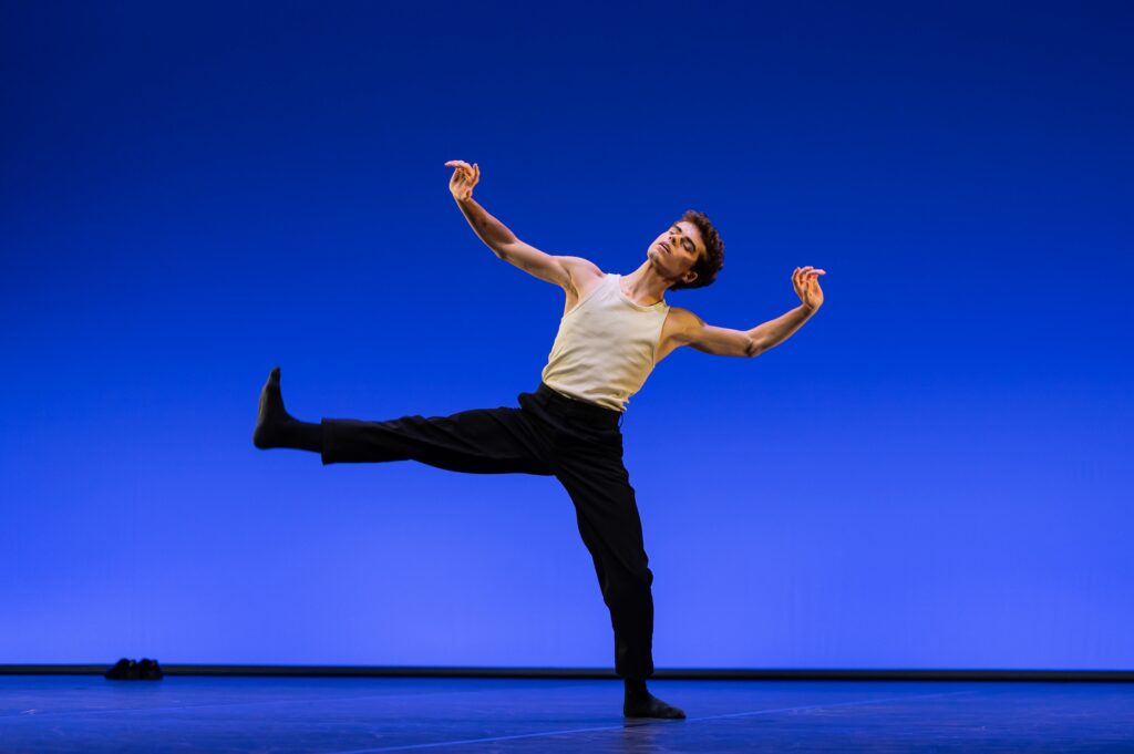 A young male dancer in a white tank top and black pants lifts his right leg up 90 degrees to the side, flexing his foot. He leans to his left slightly, closing his eyes, with his arms outstretched with flexed wrists.