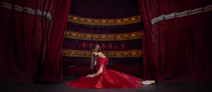Marianela Nuñez, in a bright red, full dress with tulle and ribbons, sits with one leg tucked in and one extended behind on the Royal Opera House stage. The two red curtains are partially drawn on either side of her, exposing the house behind her.