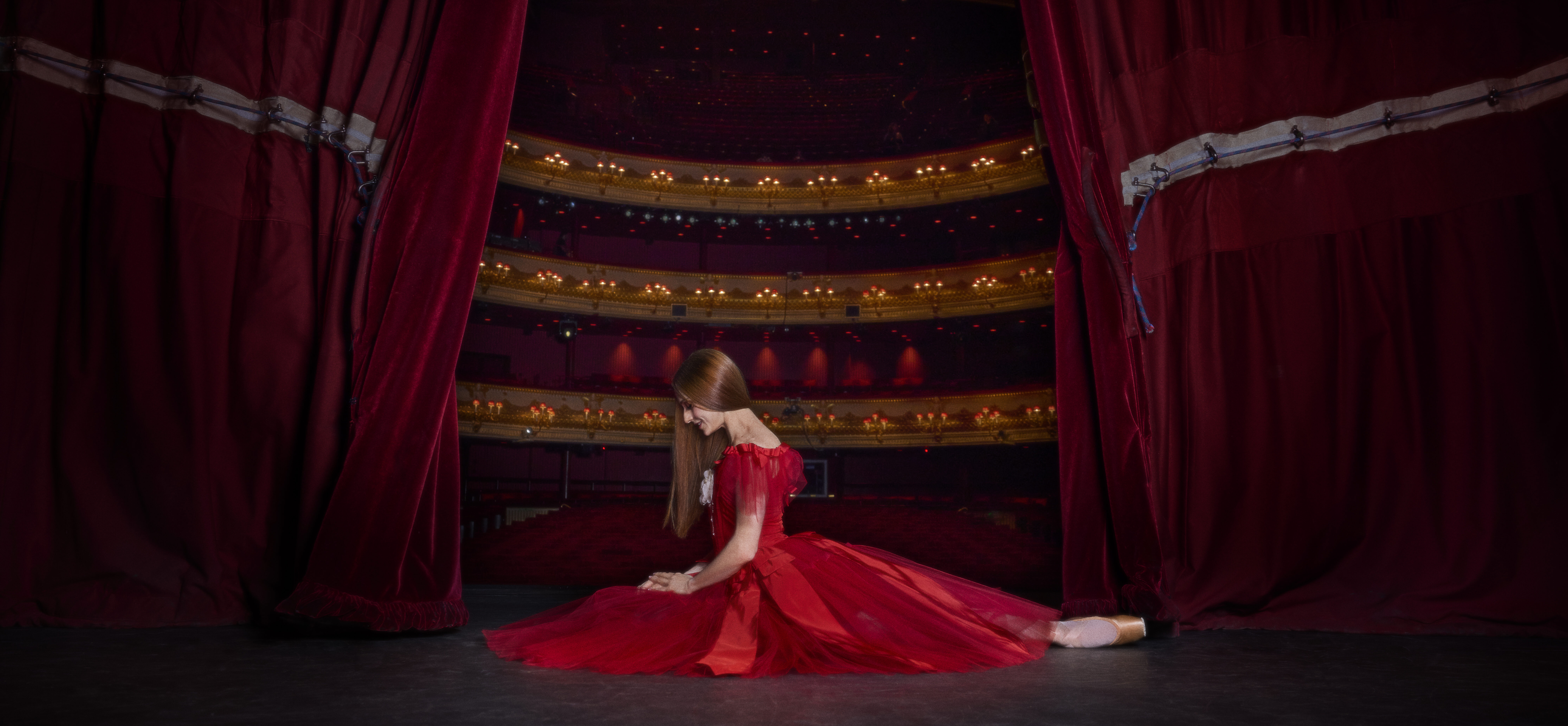 Marianela Nuñez, in a bright red, full dress with tulle and ribbons, sits with one leg tucked in and one extended behind on the Royal Opera House stage. The two red curtains are partially drawn on either side of her, exposing the house behind her.