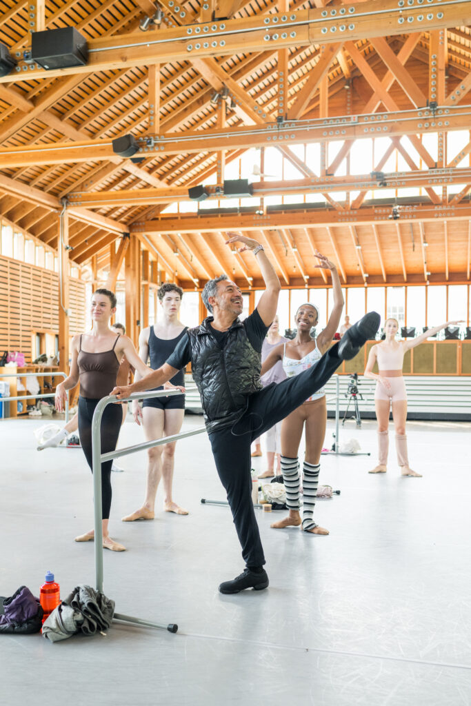 In a large indoor ballet studio with a high ceiling and wooden beams, Juan Carlos Penuela demonstrates a battement a la seconde at the barre, with one arm in high fifth. Young ballet students watch him.