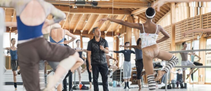 In a large indoor ballet studio with a high ceiling and wooden beams, Juan Carlos Penuela teaches a ballet class. He faces the dancers with his back to the mirror as they pose in retiré.