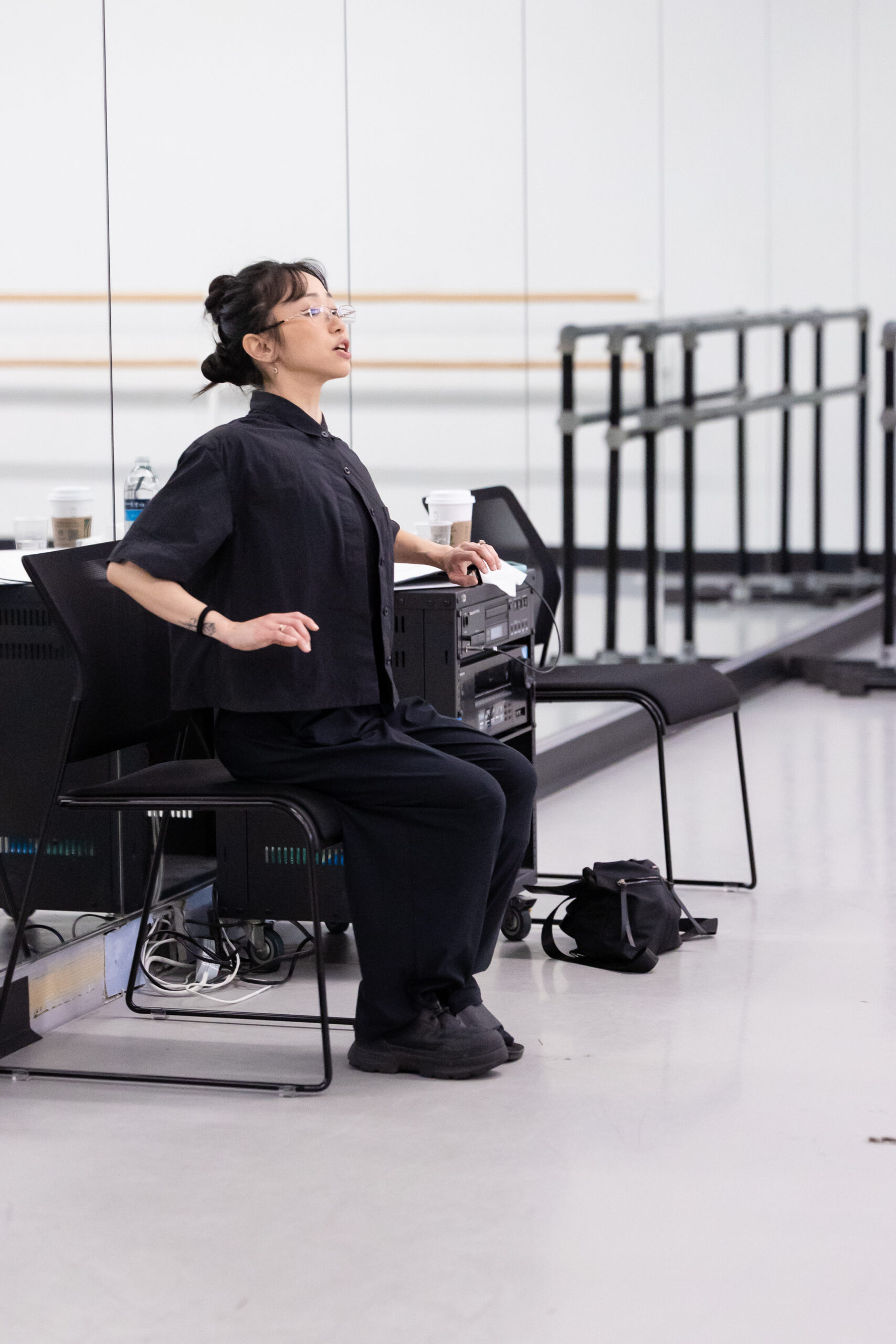 A young Asian woman sits in a chair and opens her chest, her bent and pressed back, as she watches her dance students. She wears glasses, a black shirt, black pants, and black shoes, and her hair is up.