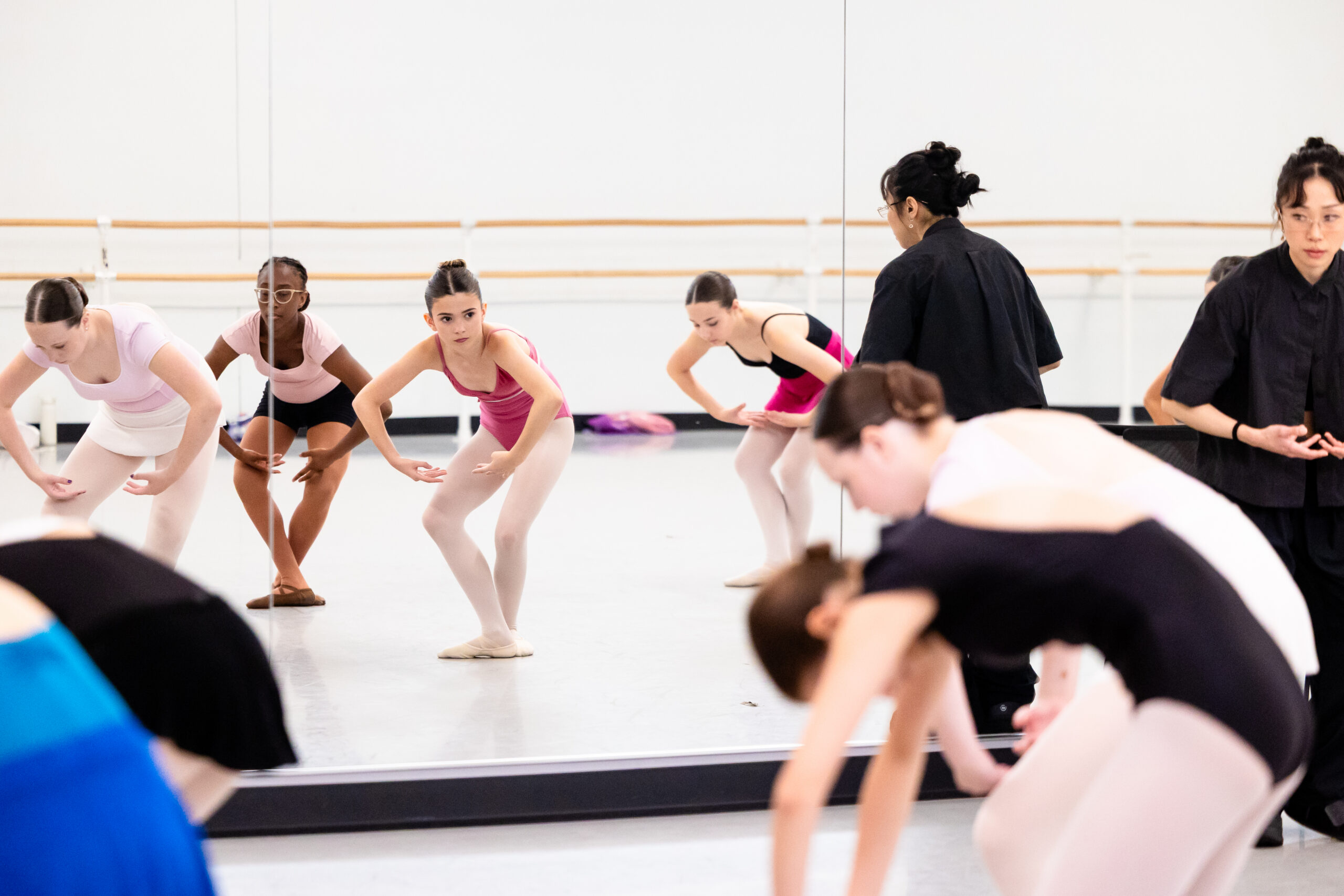 A class of young female ballet students stand in fifht position facing the left corner and contract, rounding their backs. They wear various leotards, tights, and ballet slippers. Some look up towards a teacher in the righthand corner, who is dressed in all black and watches them.