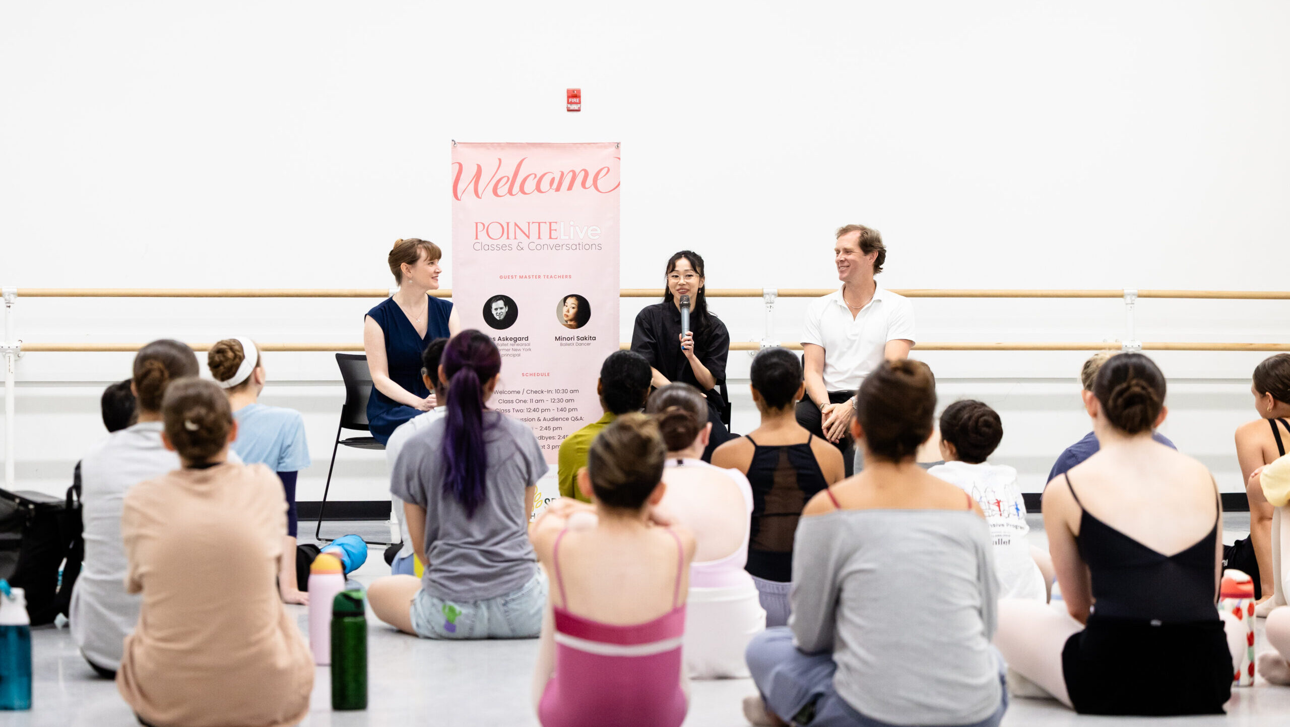 Two women and a man sit in black chairs against the wall of a large dance studio in front of a group of dancer students who wear rehearsal clothing. The woman in the middle holds a microphone and addresses the dancers.