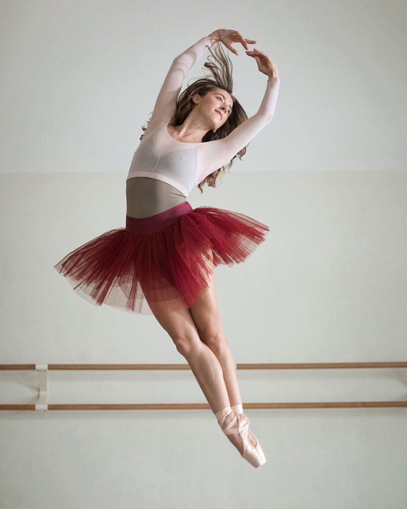 Taylor Naturkas, wearing pointe shoes, a burgundy practice tutu, brown leotard and pink shrug, jumps up wit her legs and arms in fifth position. She arches slightly to her left.