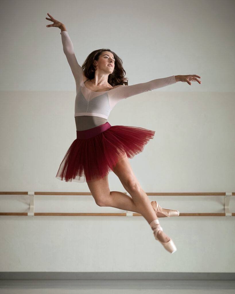 Taylor Naturkas, wearing a burgundy practice tutu, brown leotard and pink shrug, jumps up, doing a jeté. Her brown hair is long and she looks out over her left arm, which is out to the side. She lifts her right arm up.